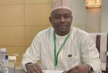 Man in white traditional attire and patterned cap sits at a conference table with a green lanyard, papers, a smartphone, and a water bottle nearby.