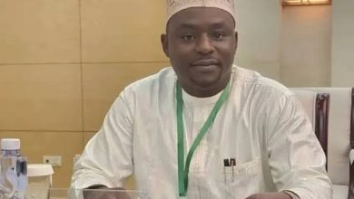 Man in white traditional attire and patterned cap sits at a conference table with a green lanyard, papers, a smartphone, and a water bottle nearby.