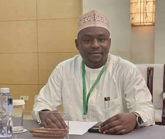 Novomed - The Times of Abuja Man in white traditional attire and patterned cap sits at a conference table with a green lanyard, papers, a smartphone, and a water bottle nearby.