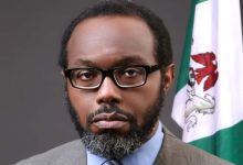 Portrait of a man with glasses and a trimmed beard wearing a brown suit and blue shirt, standing next to a Nigerian flag.