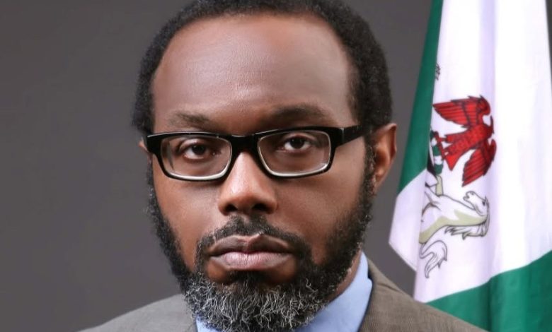 Portrait of a man with glasses and a trimmed beard wearing a brown suit and blue shirt, standing next to a Nigerian flag.