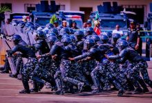 Riot police in black camouflage uniforms and helmets advance in formation with shields, as armored vehicles stand by in the background.