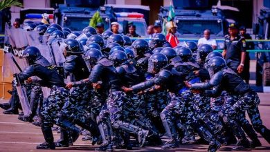 Riot police in black camouflage uniforms and helmets advance in formation with shields, as armored vehicles stand by in the background.