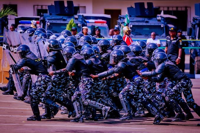 Police 1 - The Times of Abuja Riot police in black camouflage uniforms and helmets advance in formation with shields, as armored vehicles stand by in the background.