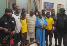 Group of people posing for a photo indoors, with two security guards on the ends and a woman in a blue dress in the center, surrounded by men and children in casual clothes.