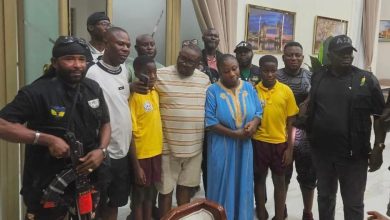 Group of people posing for a photo indoors, with two security guards on the ends and a woman in a blue dress in the center, surrounded by men and children in casual clothes.