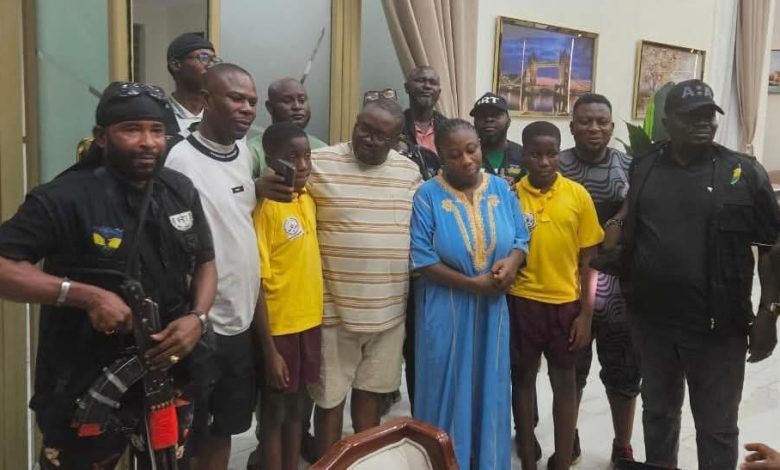 Rescue of Adelabus sister 1 - The Times of Abuja Group of people posing for a photo indoors, with two security guards on the ends and a woman in a blue dress in the center, surrounded by men and children in casual clothes.