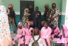 Soldiers guard a man sitting on a coffin as children and women in pink sit nearby.