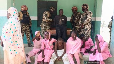 Soldiers guard a man sitting on a coffin as children and women in pink sit nearby.