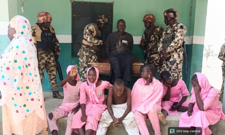 Soldiers guard a man sitting on a coffin as children and women in pink sit nearby.