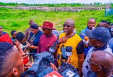 Reporters surround a man in a yellow shirt as he speaks into multiple microphones at an outdoor press conference in a grassy field, with cameras nearby.