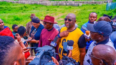 Reporters surround a man in a yellow shirt as he speaks into multiple microphones at an outdoor press conference in a grassy field, with cameras nearby.