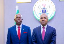 Two men in dark blue suits pose side by side in front of a round government seal on the wall, for a formal portrait.