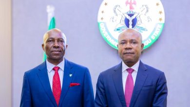 Two men in dark blue suits pose side by side in front of a round government seal on the wall, for a formal portrait.