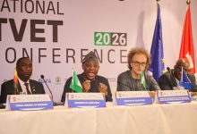 Panel of four men seated at a conference table with microphones and nameplates; flags stand behind them at a national conference backdrop reading 2026 TVET Conference/International.