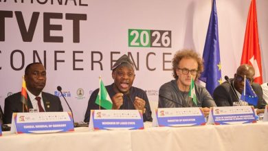 Panel of four men seated at a conference table with microphones and nameplates; flags stand behind them at a national conference backdrop reading 2026 TVET Conference/International.