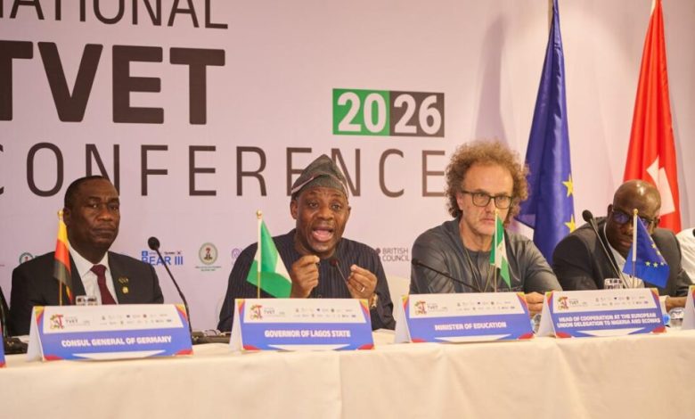 Tunji Alausa at the national TVET conference - The Times of Abuja Panel of four men seated at a conference table with microphones and nameplates; flags stand behind them at a national conference backdrop reading 2026 TVET Conference/International.