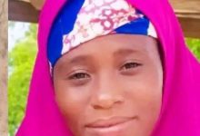 Close-up of a person wearing a bright pink hijab and a blue-pink patterned headscarf, smiling outdoors with greenery in the background.