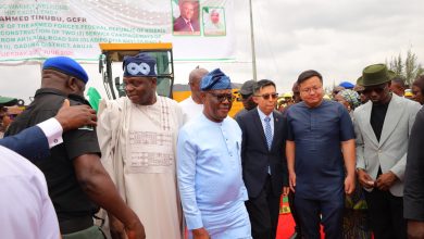 Group of men in traditional and formal attire standing on a red carpet at an outdoor event, with a large banner in the background and construction equipment nearby.