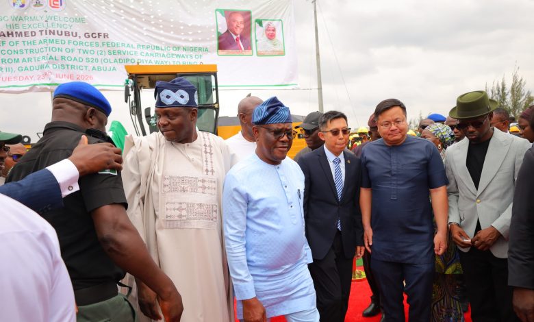 Group of men in traditional and formal attire standing on a red carpet at an outdoor event, with a large banner in the background and construction equipment nearby.
