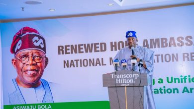 Speaker in traditional attire at a podium in front of a large political campaign banner with a portrait wearing a red cap on the left and bold text across the banner.