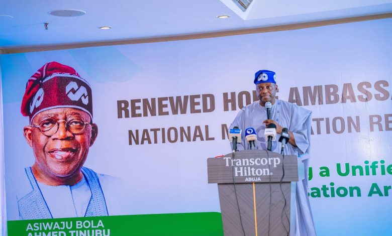 Speaker in traditional attire at a podium in front of a large political campaign banner with a portrait wearing a red cap on the left and bold text across the banner.