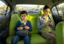 Boy in a blue blazer with a backpack sits in the bright green car back seat, frowning, while a woman beside him looks at her phone with a seatbelt fastened.