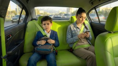Boy in a blue blazer with a backpack sits in the bright green car back seat, frowning, while a woman beside him looks at her phone with a seatbelt fastened.