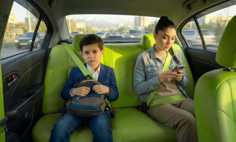Boy in a blue blazer with a backpack sits in the bright green car back seat, frowning, while a woman beside him looks at her phone with a seatbelt fastened.