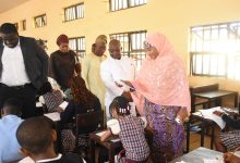 Classroom scene: students at desks writing as a woman in a pink hijab hands out papers, a man in a white suit watches nearby.