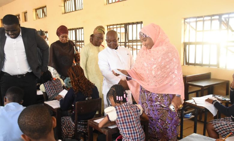 national common entrance exam - The Times of Abuja Classroom scene: students at desks writing as a woman in a pink hijab hands out papers, a man in a white suit watches nearby.