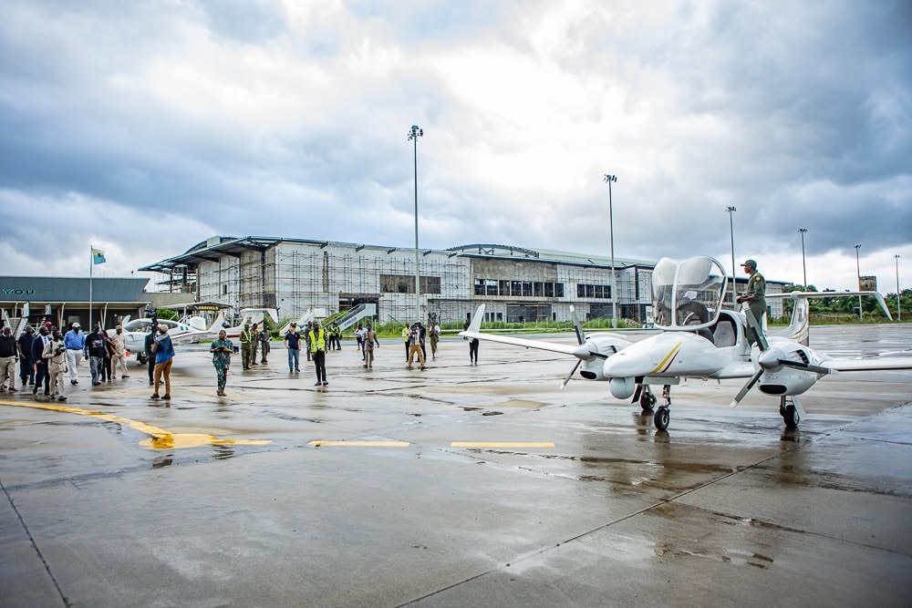 A small white airplane parked on a wet apron as onlookers stand nearby, with a construction-covered terminal building in the background under cloudy skies.