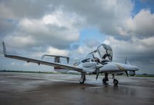 Small white twin-engine plane on a wet tarmac with open cockpit canopy and overcast sky in the background.