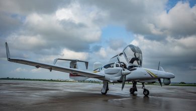 Small white twin-engine plane on a wet tarmac with open cockpit canopy and overcast sky in the background.