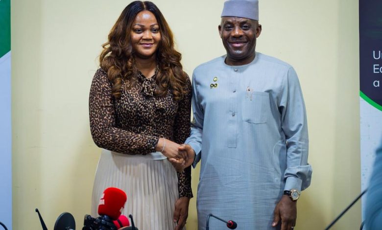 Alausa and NIMC DG - The Times of Abuja Two dignitaries shake hands at a press conference; woman in a leopard-print blouse and pleated skirt, man in gray traditional attire and cap, microphones in front.