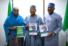 Three Nigerians in traditional attire pose together, each holding a ROOSC report on Kaduna State out-of-school children mapping, with green flags in the background.