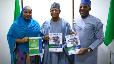 Three Nigerians in traditional attire pose together, each holding a ROOSC report on Kaduna State out-of-school children mapping, with green flags in the background.