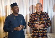 Two men stand indoors in traditional African attire, one in a dark blue outfit with a cap and the other in a colorful patterned shirt, smiling.