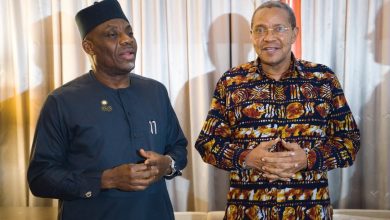 Two men stand indoors in traditional African attire, one in a dark blue outfit with a cap and the other in a colorful patterned shirt, smiling.