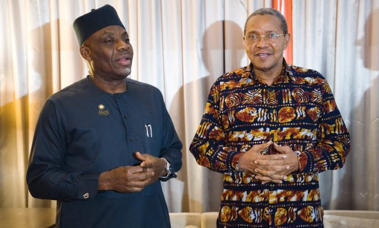 Two men stand indoors in traditional African attire, one in a dark blue outfit with a cap and the other in a colorful patterned shirt, smiling.