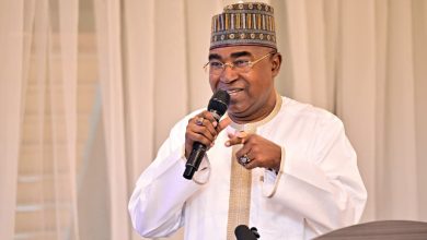 Buba Marwa - The Times of Abuja Man wearing a patterned cap and white traditional outfit speaks into a handheld microphone at a podium with a beige curtain backdrop.