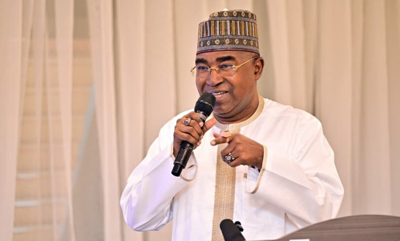 Buba Marwa - The Times of Abuja Man wearing a patterned cap and white traditional outfit speaks into a handheld microphone at a podium with a beige curtain backdrop.