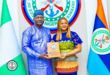 Man in blue traditional attire presents an award plaque to a woman in a colorful dress, standing before Nigeria's coat of arms and flags.