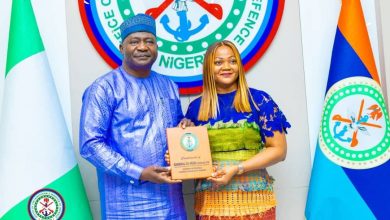 Man in blue traditional attire presents an award plaque to a woman in a colorful dress, standing before Nigeria's coat of arms and flags.