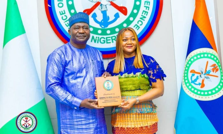 Man in blue traditional attire presents an award plaque to a woman in a colorful dress, standing before Nigeria's coat of arms and flags.