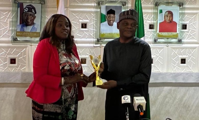 Echono receives FAN award - The Times of Abuja Two people stand at a podium exchanging a gold trophy during a formal award ceremony, with portraits on the wall behind them and microphones in front of them.