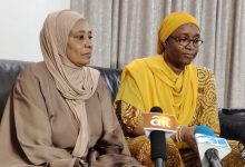 Two women sit on a sofa indoors; one in a beige hijab and matching outfit, the other in a yellow headscarf and patterned dress, speaking at a press event with microphones in front labeled 'ait' and another logo visible.