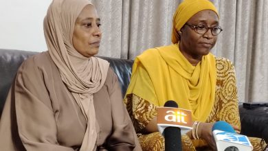 El-Rufais wife - The Times of Abuja Two women sit on a sofa indoors; one in a beige hijab and matching outfit, the other in a yellow headscarf and patterned dress, speaking at a press event with microphones in front labeled 'ait' and another logo visible.