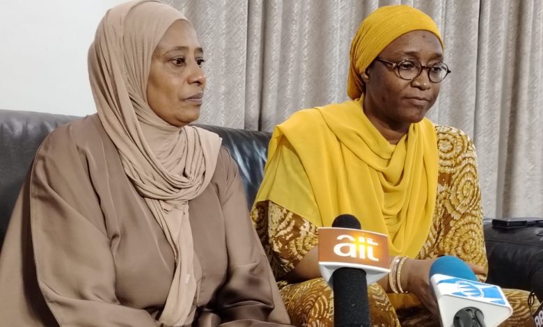 El-Rufais wife - The Times of Abuja Two women sit on a sofa indoors; one in a beige hijab and matching outfit, the other in a yellow headscarf and patterned dress, speaking at a press event with microphones in front labeled 'ait' and another logo visible.