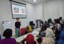 Instructor standing at the front of a classroom giving a presentation to a diverse group seated around tables with a projected slide behind her